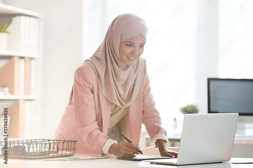 Poster positive young muslim designer in hijab standing at table and editing digital sketch using drawing p