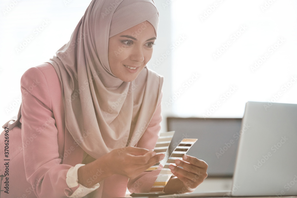 Poster content attractive arabian designer in pink hijab leaning on table and comparing color palettes whil