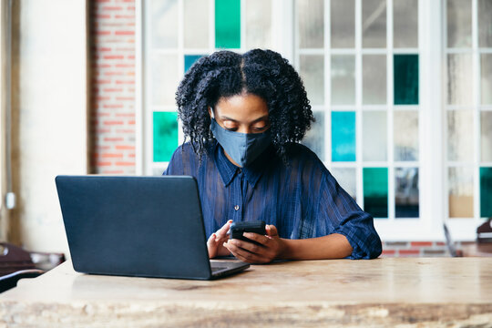 Black woman working at cafe on laptop and phone with face mask during covid-19