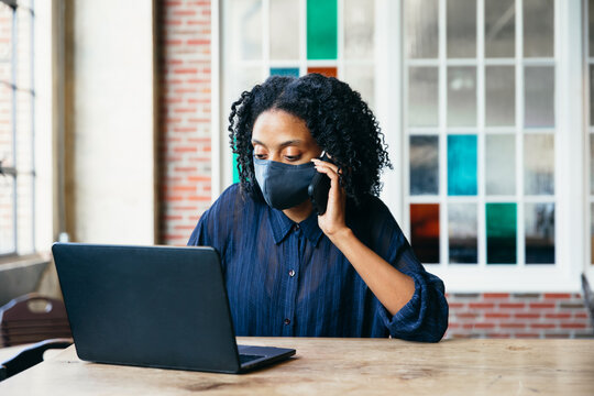 Black woman working at cafe on laptop and phone with face mask during covid-19