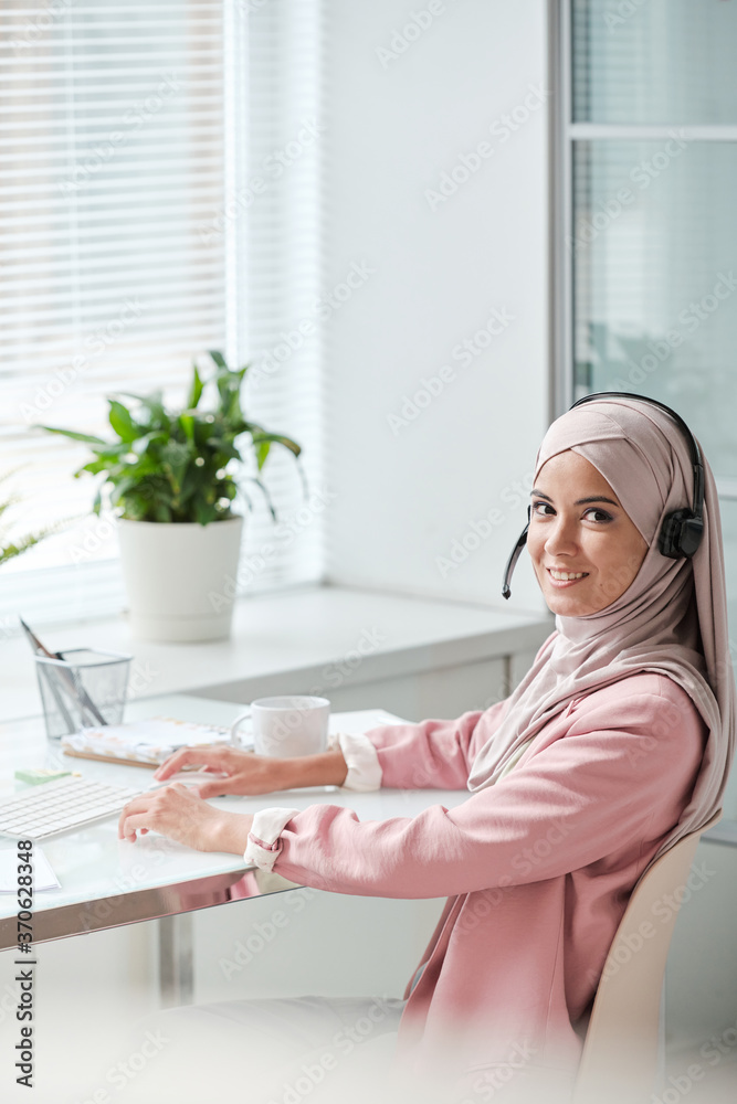 Poster portrait of smiling young muslim call center operator in traditional veil sitting at desk and talkin