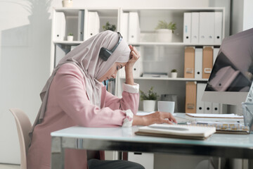 Obraz na płótnie Canvas Young Arabian businesswoman in hijab tired from work in call center sitting at desk and leaning head on hand