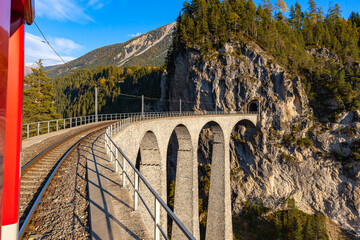 Reis met de rode Rhätische Bahn-sightseeingtrein Bernina Express die op zonnige herfstdag over het Landwasser-viaduct rijdt met blauwe luchtwolk, Canon van Graubünden, Zwitserland
