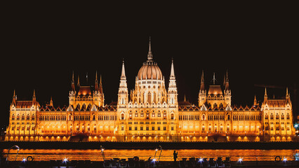 Fototapeta na wymiar Panoramic background orange night image of Budapest parlament from the front side with boat and tourist taking a photo of the city. Travel in hungary and historical sites.