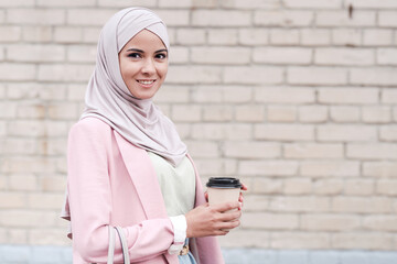 Portrait of positive beautiful young Muslim woman in hijab standing against brick wall and drinking coffee on move