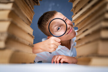 Fototapeta premium Schoolboy sitting between two piles of books looking at camera through the magnifying glass.