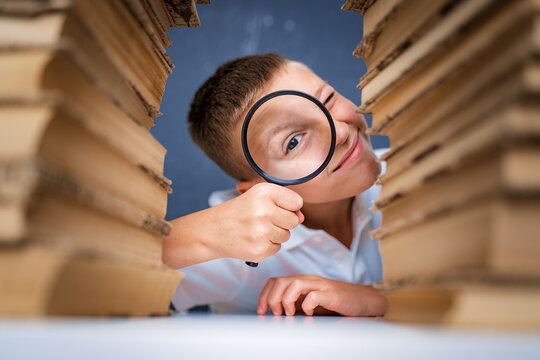 Schoolboy sitting between two piles of books looking at camera through the magnifying glass.