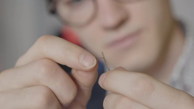 Extreme close-up of male hands threading needle with blurred face of tailor at the background. Portrait of Caucasian dressmaker in eyeglasses working in atelier. Serious designer sewing garment.