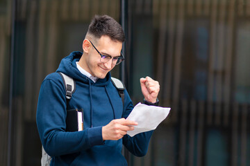 Happy positive successful college student, cheerful pupil, young handsome man with book smiling in glasses, looking at result list. Smart boy passed university exam, test, celebrating victory, triumph