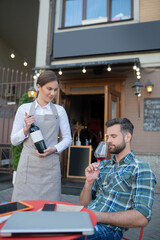 Bearded male smelling red wine, smiling waitress showing him wine bottle