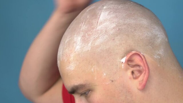 The bald young man insolently shaves his head closeup. A man cuts his hair with a razor on a blue background.