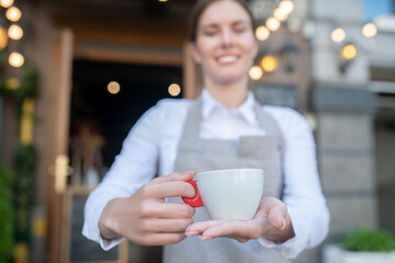 Cute waitress in grey apron offering cup of coffee