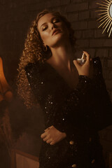 Beautiful woman uses perfume fragrance. Model wearing trendy sequin dress, posing in dark vintage...
