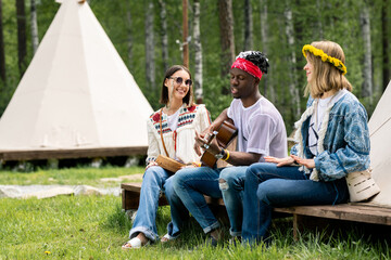 Young Black guy in bandana sitting with girls at tent and singing beautiful song at campsite