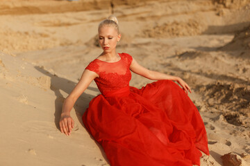 Attractive blonde woman in a red dress lies on the sand, posing in the desert.