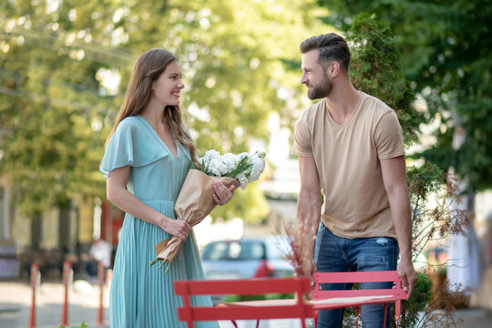 Bearded male pulling chair for cute young female with bouquet of flowers
