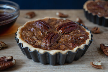 Head on view of a individual size pecan pie tart in case.  Pecans and caramel sauce seen at edges of frame against a dark background and a natural light wood surface