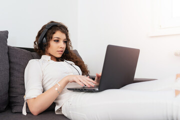 Pretty girl in white clothes lying on the couch, wearing headphones and looking at laptop.