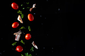 Falling pieces of fresh bacon, cherry tomatoes, parsley and onions on a black background, freeze in motion, for design, cooking