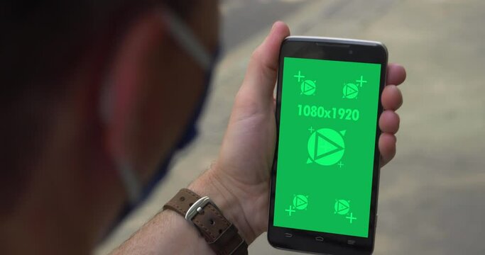 An over-the-shoulder shot of a man in a face mask using or talking on a green screen smartphone. Optional tracking points for screen replacement.  	