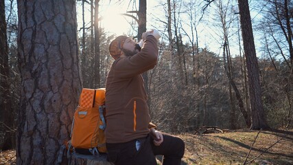 A young tired Caucasian man carries a rest break with a backpack sitting and drinking water on a tree in a nature forest during hikes, trips, vacations, on vacation, outdoor lifestyle freedom concept