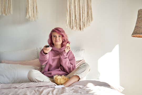 Smiling hipster gen z teen girl with pink hair watching tv movie series sitting in bed, holding remote control, eating chips snack. Teenager relaxing watching tv television reality show in bedroom.