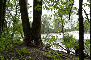 Redhead woman sitting in the middle of a forest seeing her camera next to a huge log on the shore of a lake wearing a red hat