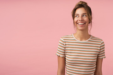 Portrait of young charming brunette female with natural makeup smiling widely while looking cheerfully aside, standing over pink background with hands down