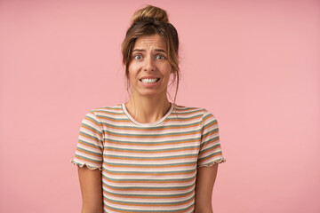 Studio photo of young green-eyed brunette female showing her teeth while grimacing confusedly face, isolated against pink background with hands down
