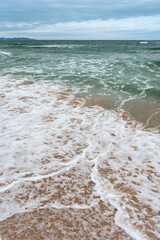Fototapeta na wymiar Turquoise sea with waves and sea foam, beach, cloudy sky