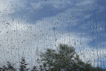 Drops of water from heavy rain run down the dirty glass, which reflects the blue cloudy sky.