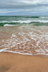Fototapeta na wymiar Turquoise sea with waves and sea foam, sandy beach, cloudy sky