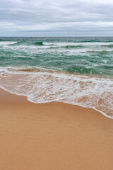 Turquoise sea with waves and sea foam, sandy beach, cloudy sky