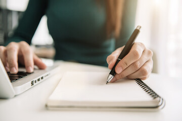 Female businessman hand working at a computer and writing on a notepad with a pen in the office. 