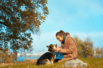 Autumn pet walking. woman in stylish coat sits with her Welsh Corgi dog in autumn park, looks at each other, eye contact with pet. Concept friendship with dog and human, autumn portrait