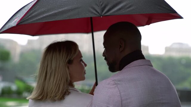 multiracial loving couple is standing under umbrella in rainy day and kissing