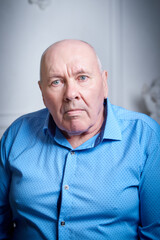Portrait of an elderly bald man in a blue shirt in the room. A poor pensioner lives alone.