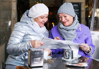 Fototapeta na wymiar Two positive mature women tourists with city guide sitting at terrace cafe
