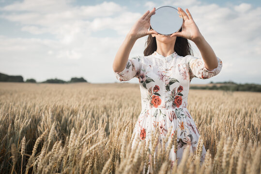 Young, pretty girl with long hair in a light dress with a floral print, holding a round mirror in a wheat field, posing in front of the camera, in the background of the sunset, nature, pure ecology