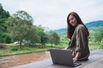 Portrait image of a beautiful asian woman working on laptop computer while sitting by the river with mountains and nature background