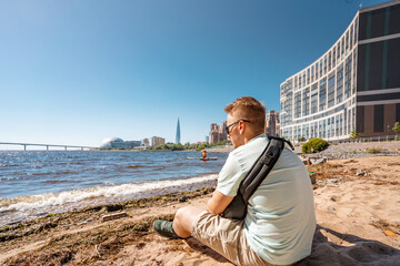Fototapeta na wymiar A young man with a backpack made a stop on the beach of Saint Petersburg on the Gulf of Finland with a view of business centers and the Lakhta Center skyscraper