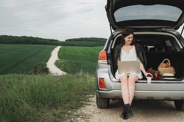 Young Stylish Woman Sits on the Trunk of Her SUV Car and Working on a Laptop in the Green Field