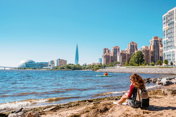 A girl sits on the beach by the sea of the Gulf of Finland in St. Petersburg with a view of the Lakhta Center skyscraper