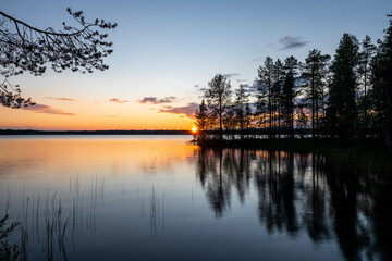 Beautiful summer sunset over finnish lake with silhouette of trees reflecting in water in gold, orange and blue, Finland, Kuusamo, Finland