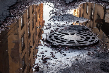 Sewer manhole in a muddy puddle with reflections of old houses