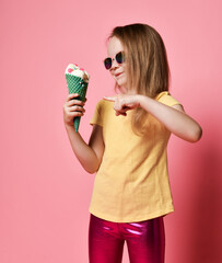 Sly smiling kid girl in t-shirt and leggings is pointing at big vanilla ice cream with candies in waffles cone she holds