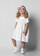 Cute smiling blonde curly kid girl princess in white casual dress and sneakers is standing walking over grey wall