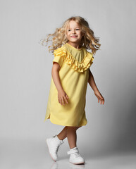 Frolic smiling blonde curly kid girl in yellow dress and sneakers walking moving over grey wall background.