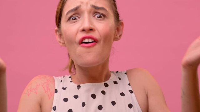 Close up portrait of an angry caucasian female with black tied hair shouting WTF and grabbing her hands behind her head on a pink background. Blaming, warning, accusing concept.