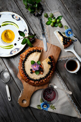 Big round cheese cake with black currant and a cup of coffee on a wooden grey table.